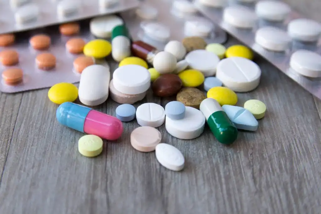 A variety of colorful pills, capsules, and tablets are scattered on a wooden surface, with several blister packs of medication in the background.