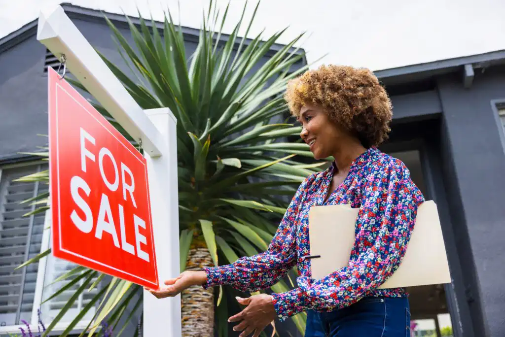 A woman stands outside a house, smiling and holding documents, next to a large red For Sale sign. The house and a tall plant are visible in the background.