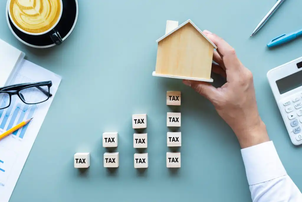 A hand holds a small wooden house above stacked wooden blocks labeled TAX, forming a staircase. Nearby are a calculator, eyeglasses, a coffee cup, and financial charts on a light blue table.