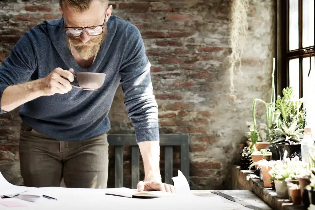 A bearded man wearing glasses and a blue sweater leans over a desk, holding a mug, in a sunlit room with a rustic brick wall and several potted plants on the windowsill.