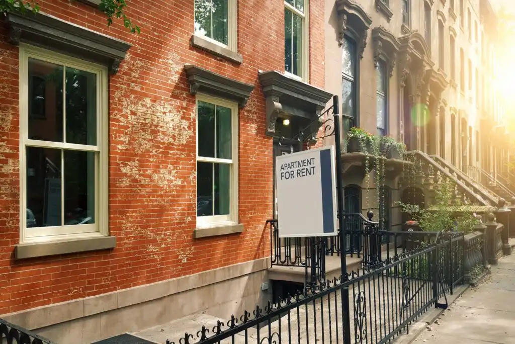 A For Rent sign hangs outside a red brick apartment building with large windows and iron railings, on a sunlit city street lined with similar historic buildings.