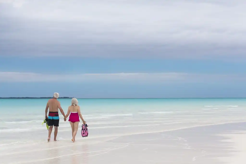 An older couple holding hands walks along a serene beach shoreline, carrying snorkeling gear, with calm turquoise water and a cloudy sky in the background.
