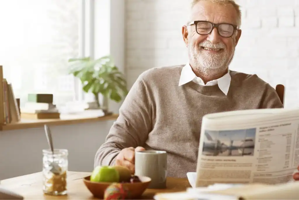 Smiling older man with glasses sits at a table, holding a mug and reading a newspaper. A fruit bowl and an open jar are on the table. Sunlight streams in through a window, creating a cozy atmosphere.