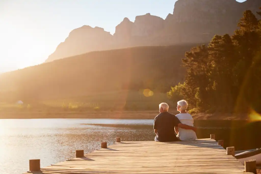 An elderly couple sits closely together on a wooden dock overlooking a calm lake, surrounded by trees and mountains, with warm sunlight illuminating the scene.