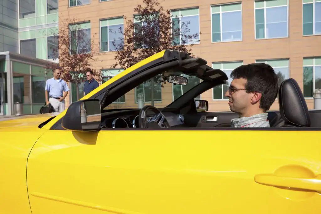 A man sits in a yellow convertible car with the top down, parked in front of a modern office building. Two men are walking on the sidewalk nearby, and trees are visible in the background.