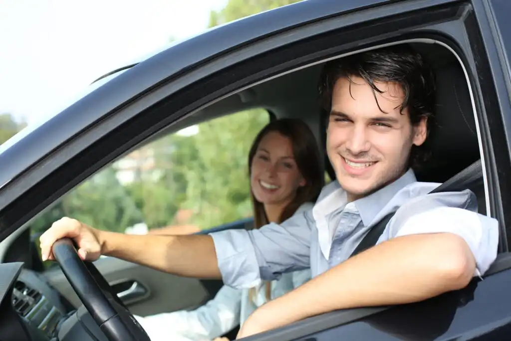 A young man sits in the driver’s seat of a car, smiling, with a young woman in the passenger seat beside him. Both appear happy and relaxed, and the car window is open with greenery visible outside.