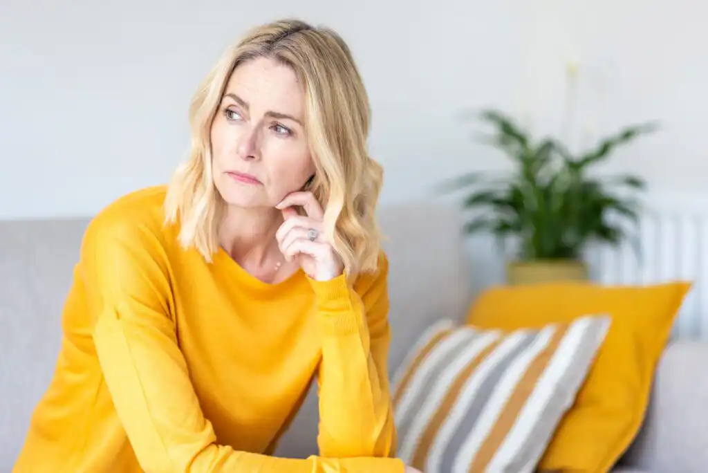 A woman with blonde hair wearing a yellow sweater sits indoors, looking to the side with a thoughtful expression. Behind her are a striped cushion and a green potted plant on a light-colored couch.