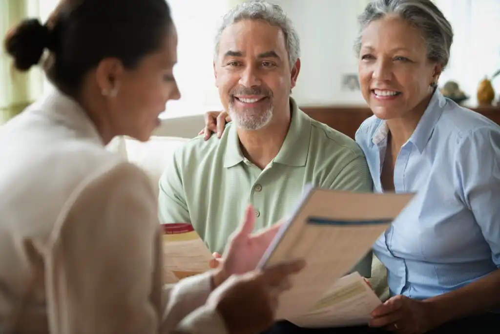 An older couple sits together, smiling and listening to a woman holding documents during a meeting at home. The couple appears engaged and attentive as they review paperwork with the advisor.