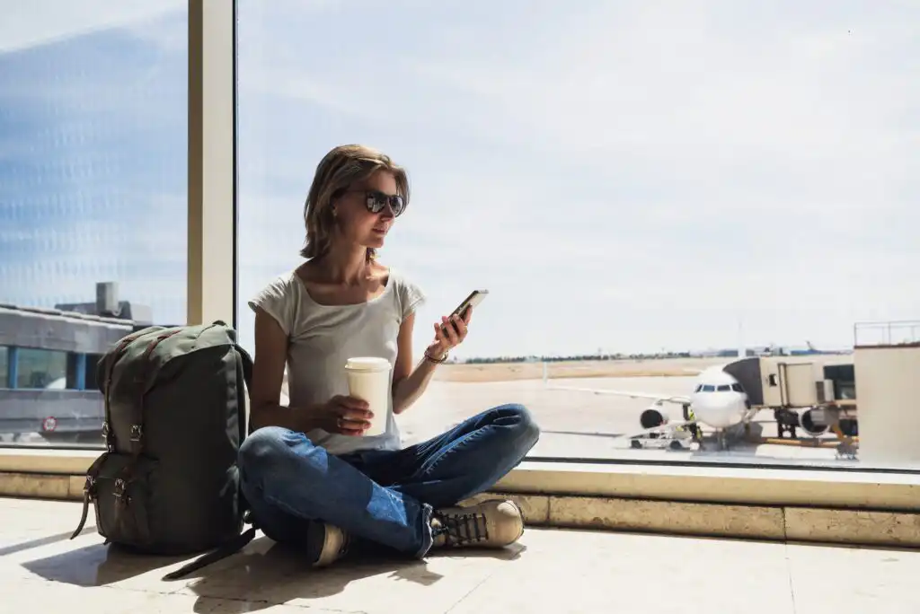 A woman wearing sunglasses sits cross-legged on the floor by an airport window, holding a coffee cup and looking at her phone. A large backpack is next to her, and airplanes are visible outside.