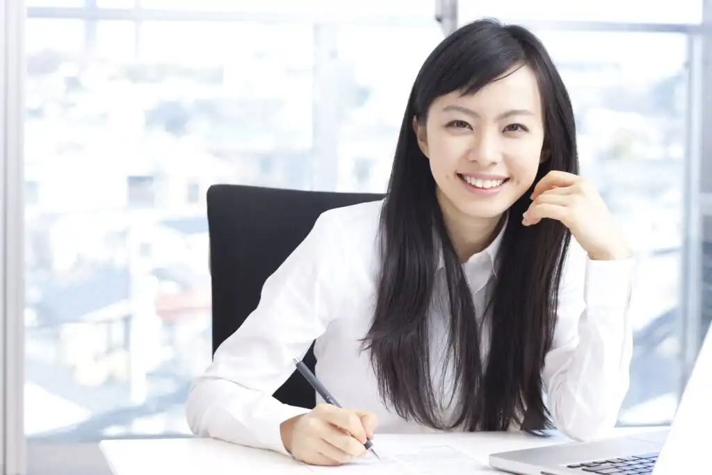 A woman with long black hair, wearing a white shirt, sits at a desk with papers and a laptop, smiling at the camera while holding a pen. A bright window is in the background.
