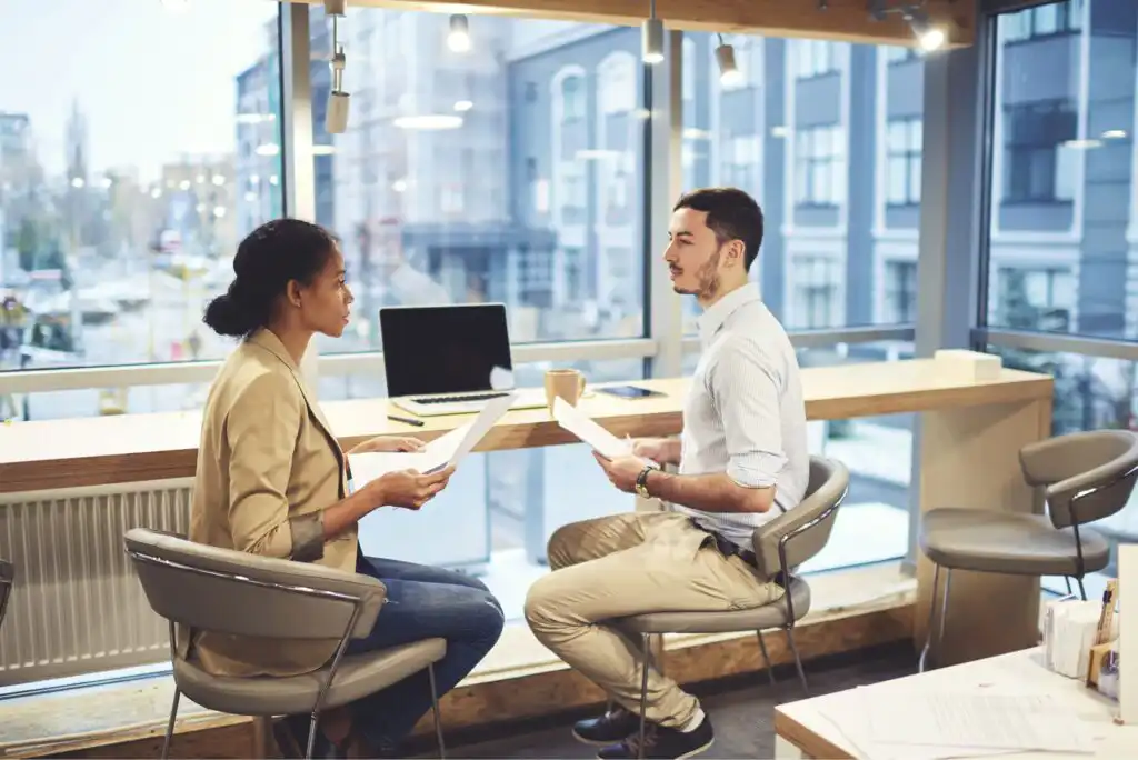Two people sit across from each other in a modern office, holding papers and having a discussion near a large window with city views. A laptop and other work materials are on the counter beside them.