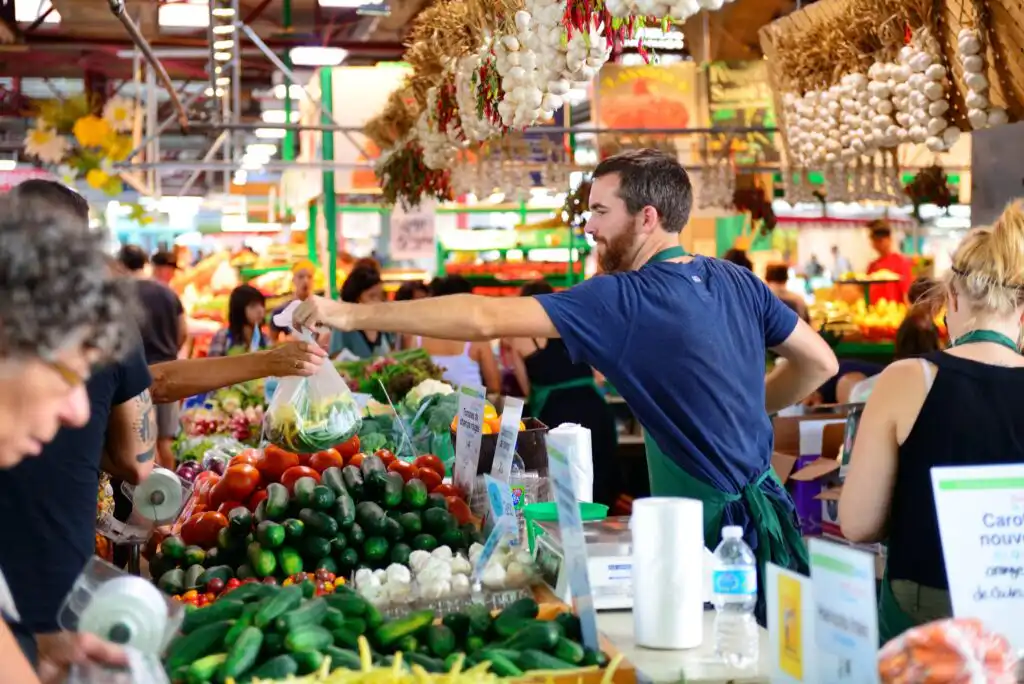 A bustling indoor farmers market scene with vendors and customers exchanging goods. Fresh vegetables, tomatoes, cucumbers, and garlic hang overhead. People are shopping and interacting at colorful produce stalls.