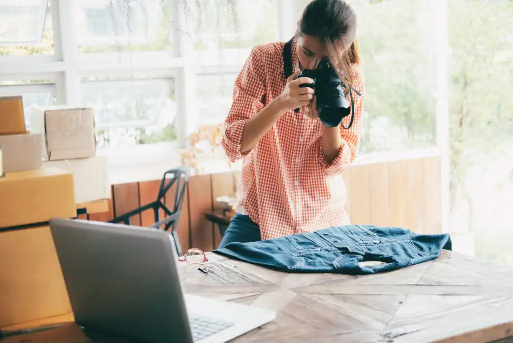 A person wearing a checked shirt takes a photo of a denim jacket laid on a wooden table, with a laptop and cardboard boxes nearby in a bright room.