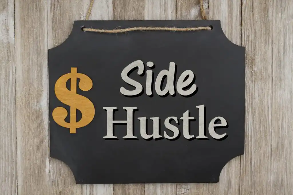 A black sign with a rope hanger displays a gold dollar sign and the words Side Hustle in large, bold letters, hanging against a wooden background.