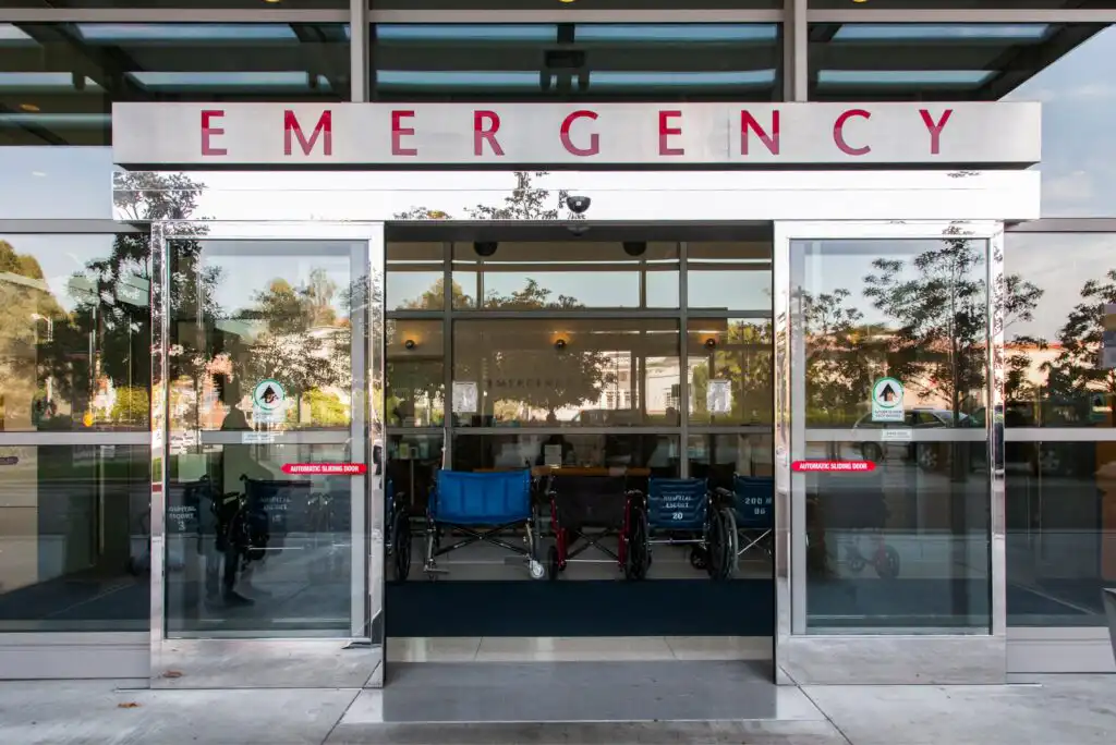 Entrance to a hospital emergency room with automatic glass doors. Wheelchairs are lined up inside, and the word “EMERGENCY” is prominently displayed above the entrance.