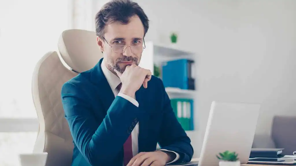 A man in a suit and glasses sits at a desk with a laptop, resting his chin on his hand and looking thoughtfully at the camera in a modern office setting.