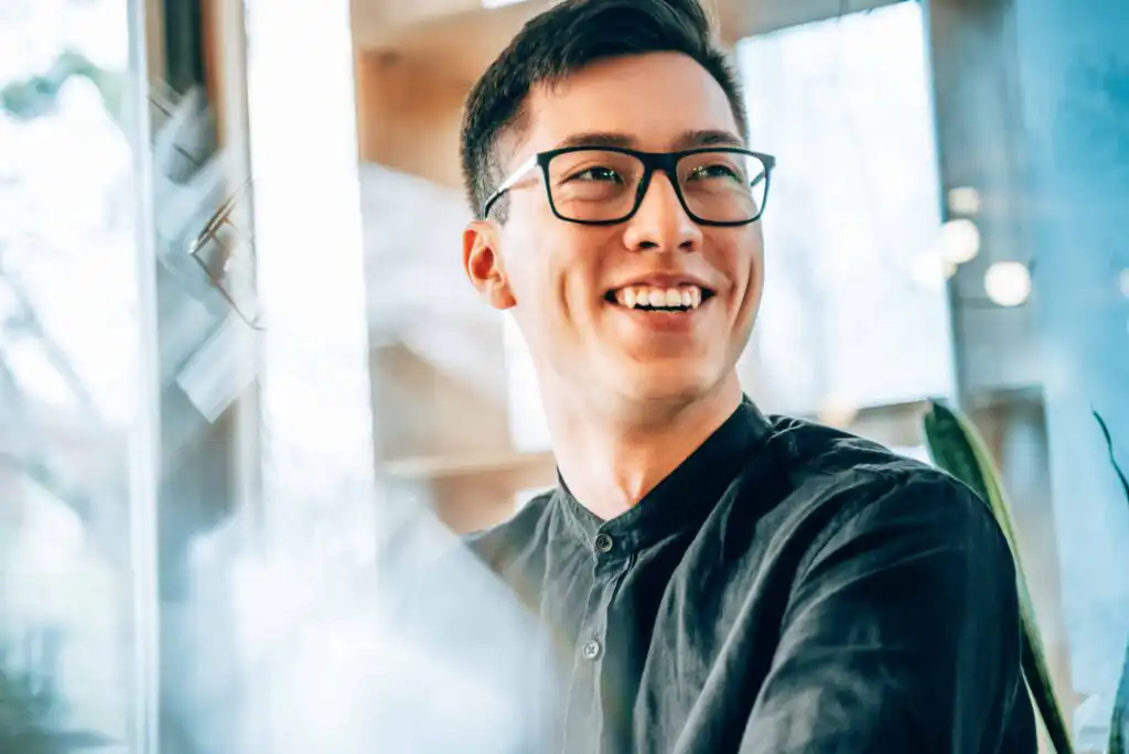 A young man wearing glasses and a dark shirt smiles while sitting indoors near a window, with soft natural light illuminating his face.