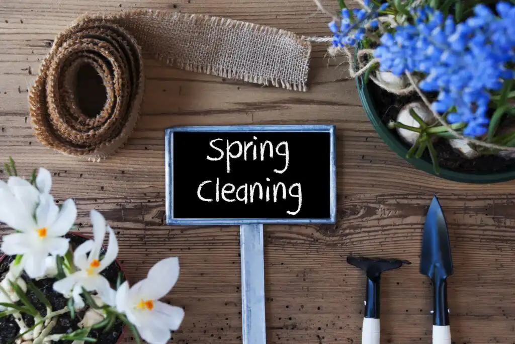 A small chalkboard sign with Spring Cleaning written on it is surrounded by gardening tools, burlap ribbon, and flowering white and blue plants on a wooden table.