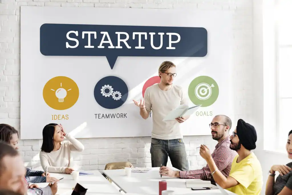 A group of people sit around a table in a meeting room, listening to a man standing and speaking. Behind him is a banner that reads STARTUP with icons labeled Ideas, Teamwork, and Goals.