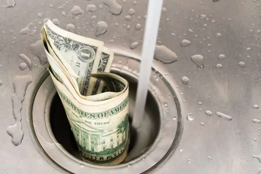 Rolled-up U.S. dollar bills being soaked under running water in a metal sink, with water droplets nearby, symbolizing wasting or washing away money.