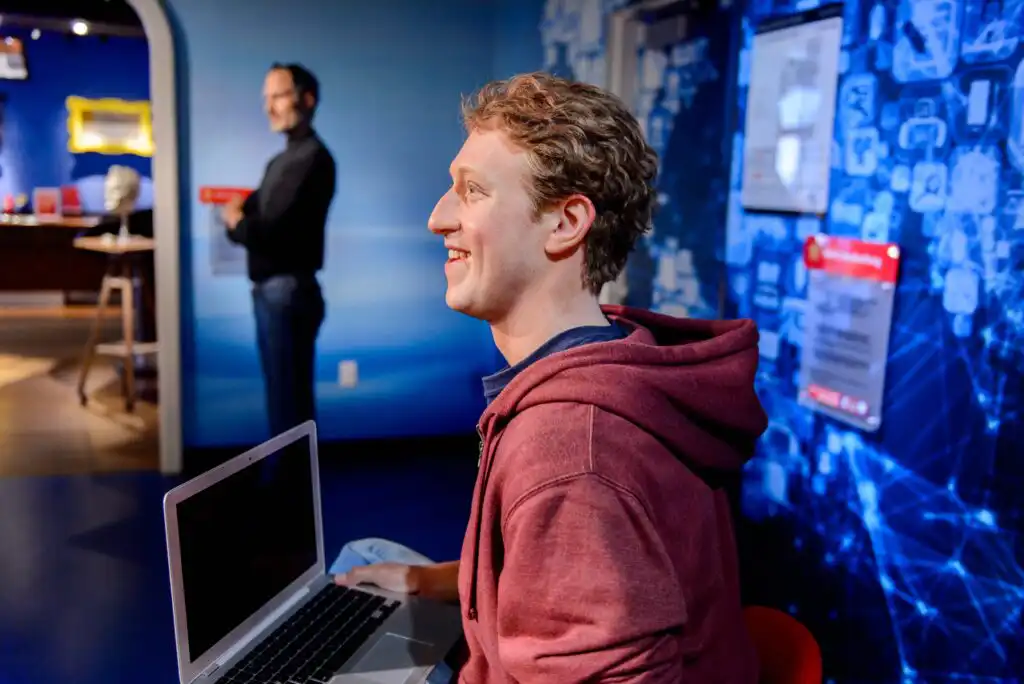 A wax figure of a smiling young man in a red hoodie sits holding a laptop in a museum display with a blue tech-themed background; another figure stands in the background.