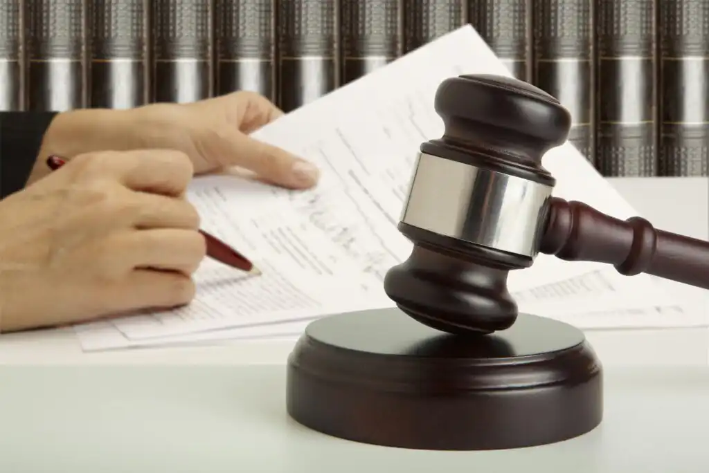 A wooden judge’s gavel rests on a sound block in the foreground, while a person reviews and marks documents with a pen in the background, with shelves of law books behind them.