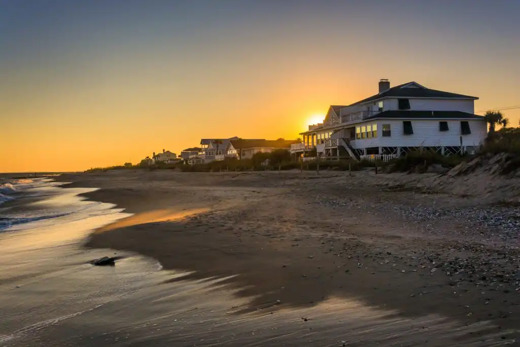 Houses line a quiet sandy beach at sunset, with the sun glowing behind the buildings and gentle waves washing up on the shore.