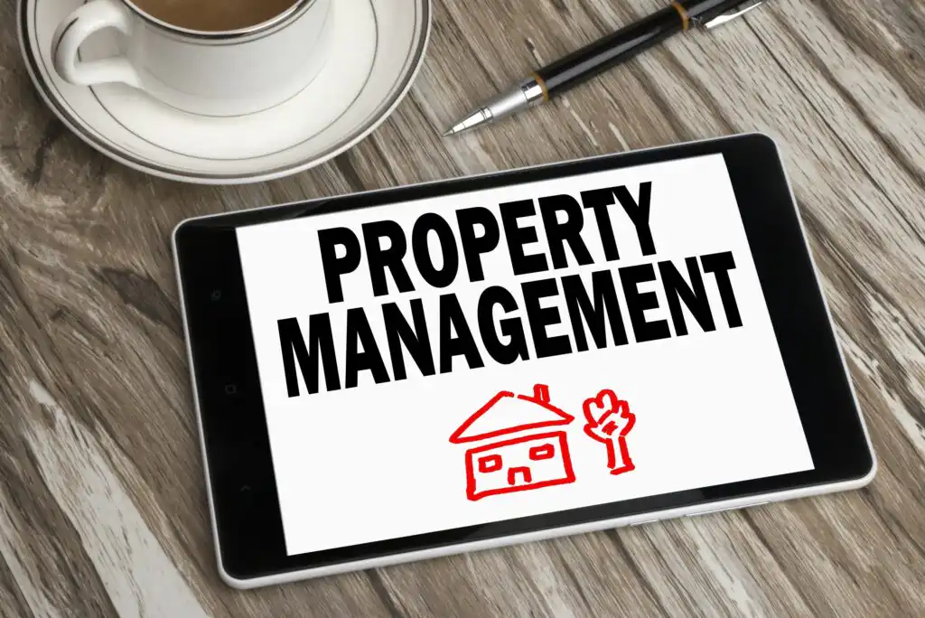 A tablet on a wooden table displays the words Property Management above a red drawing of a house and tree. A cup of coffee and a pen are placed nearby.