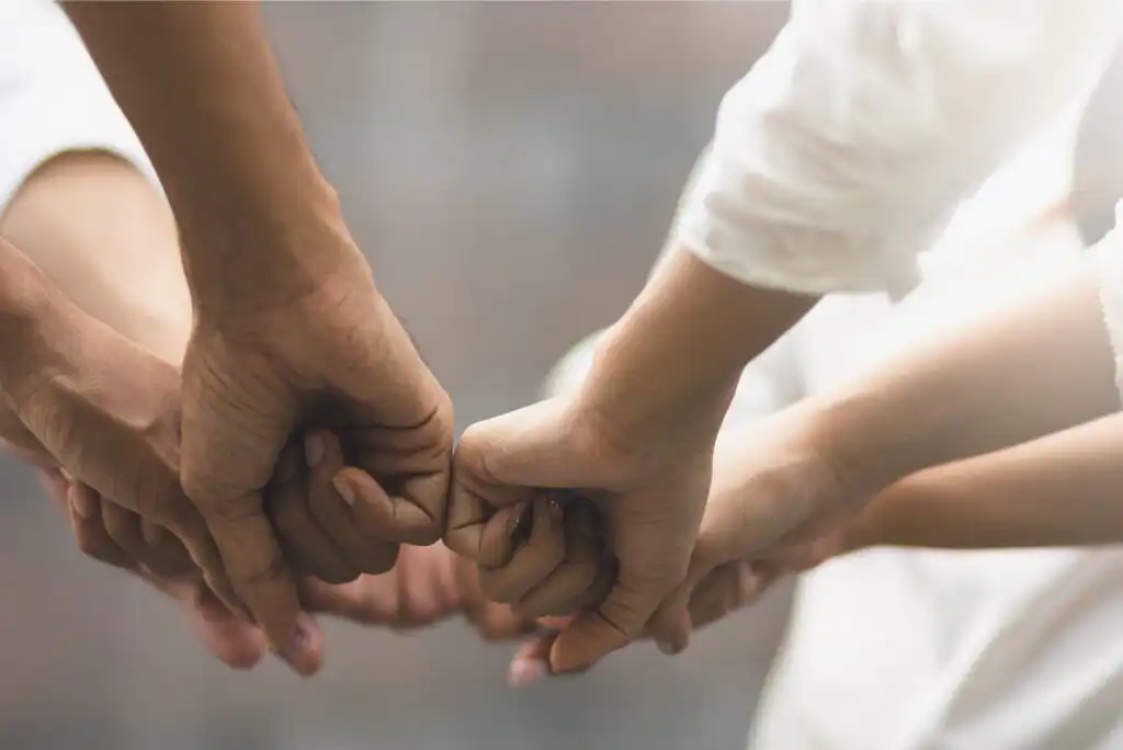 Five hands coming together in a group fist bump, symbolizing unity, teamwork, and support. The background is blurred, and the people are wearing white or light-colored clothing.