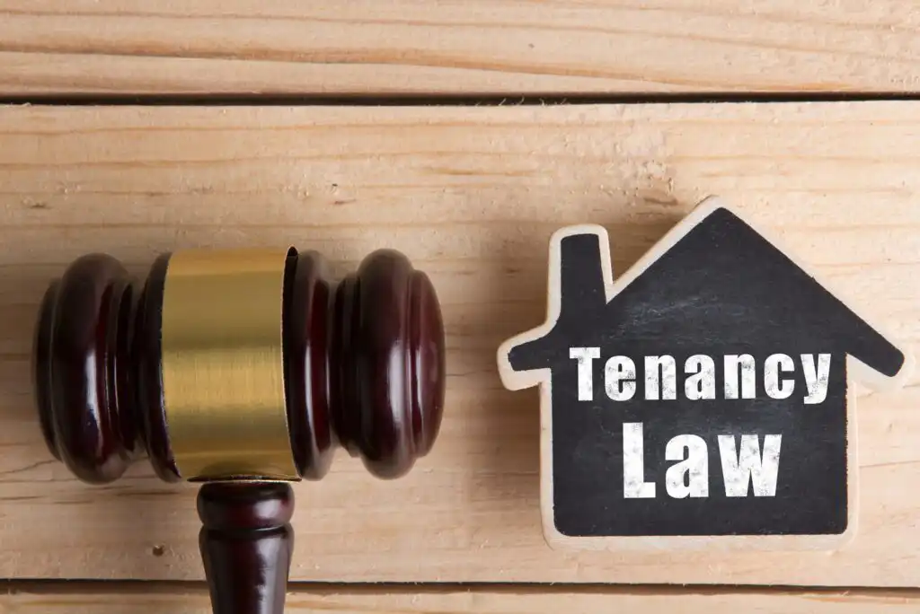 A wooden judges gavel rests next to a black house-shaped sign with the words Tenancy Law written on it, all placed on a light wooden surface.