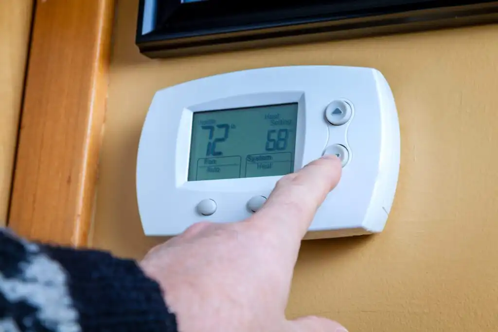A person adjusts a digital thermostat mounted on a yellow wall. The display shows an indoor temperature of 72°F and a heat setting of 68°F. The persons finger is pressing an arrow button on the device.