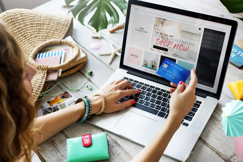 A person shops online using a laptop, holding a credit card in one hand and typing with the other. The table has a straw bag, notebook, pen, wallet, and tropical leaves, creating a summery atmosphere.