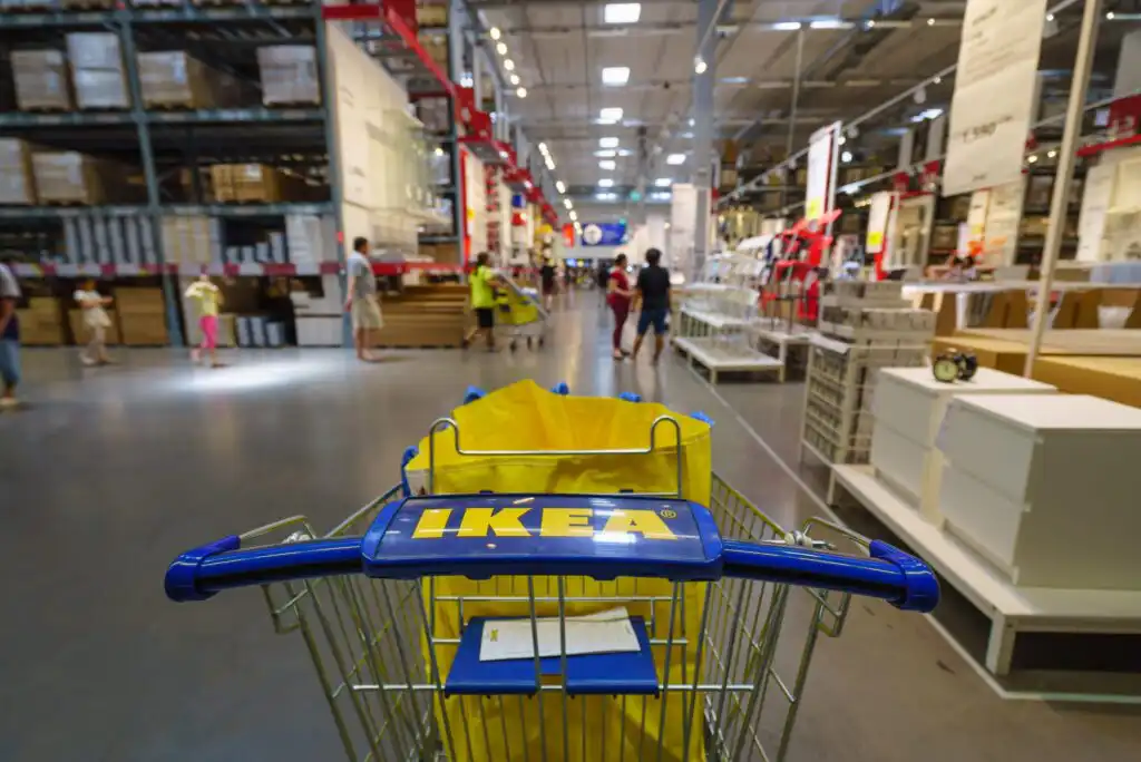 A shopping cart with an IKEA logo is pushed down an aisle inside a spacious IKEA store, with shelves of furniture, home goods, and several shoppers visible in the background.