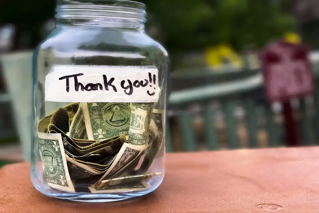 A glass jar labeled Thank you!! with a smiley face, partially filled with folded U.S. dollar bills, sits on a surface outdoors with a blurry green background.