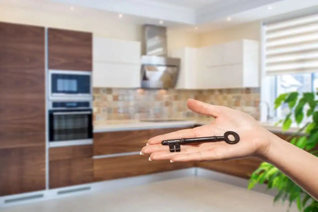 A hand holding a large vintage key in the foreground, with a modern kitchen featuring wood cabinets, built-in appliances, and tiled backsplash in the background.