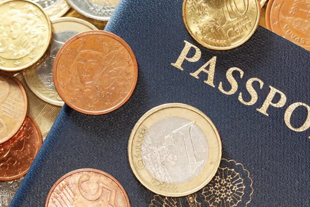 A close-up of various Euro coins scattered on top of a dark blue passport, with the word PASSPORT partially visible in gold letters.