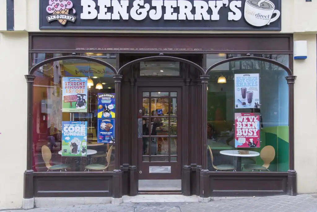 The exterior of a Ben & Jerry’s ice cream shop with large windows displaying posters about student discounts, sundaes, milkshakes, and special offers; inside are empty tables and chairs.