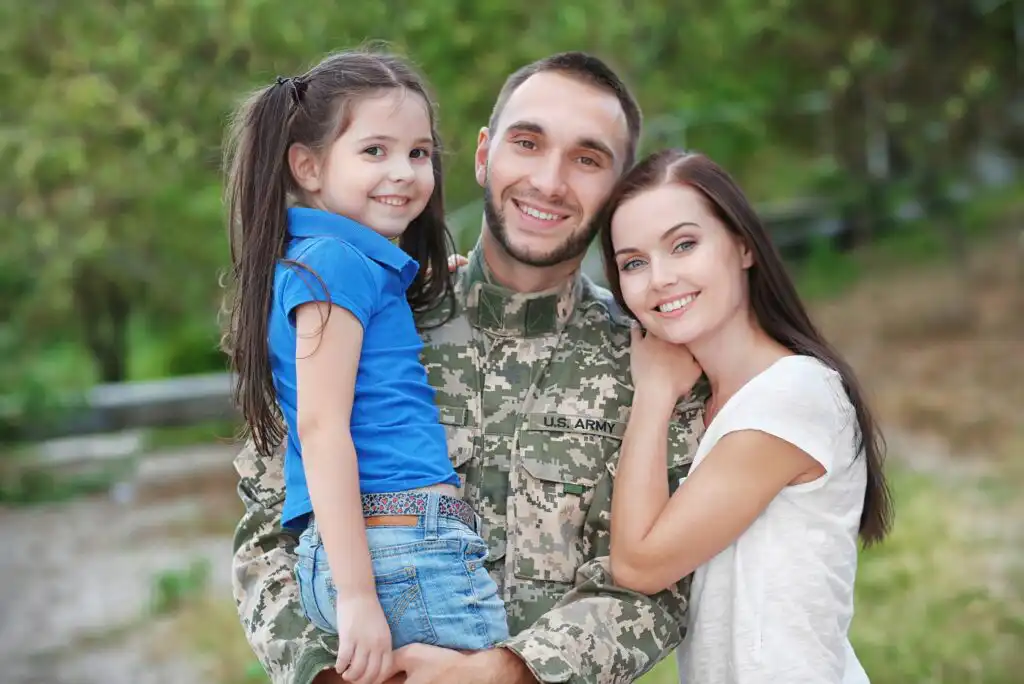 A man in a U.S. Army uniform smiles while holding a young girl, with a woman standing close beside them. They are outdoors, appearing happy and affectionate.