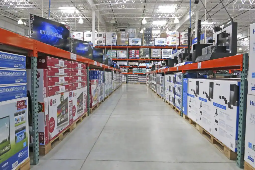 A warehouse store aisle with large TVs and electronics stacked on pallets, bright overhead lighting, and shelves stocked high with boxed products on both sides.
