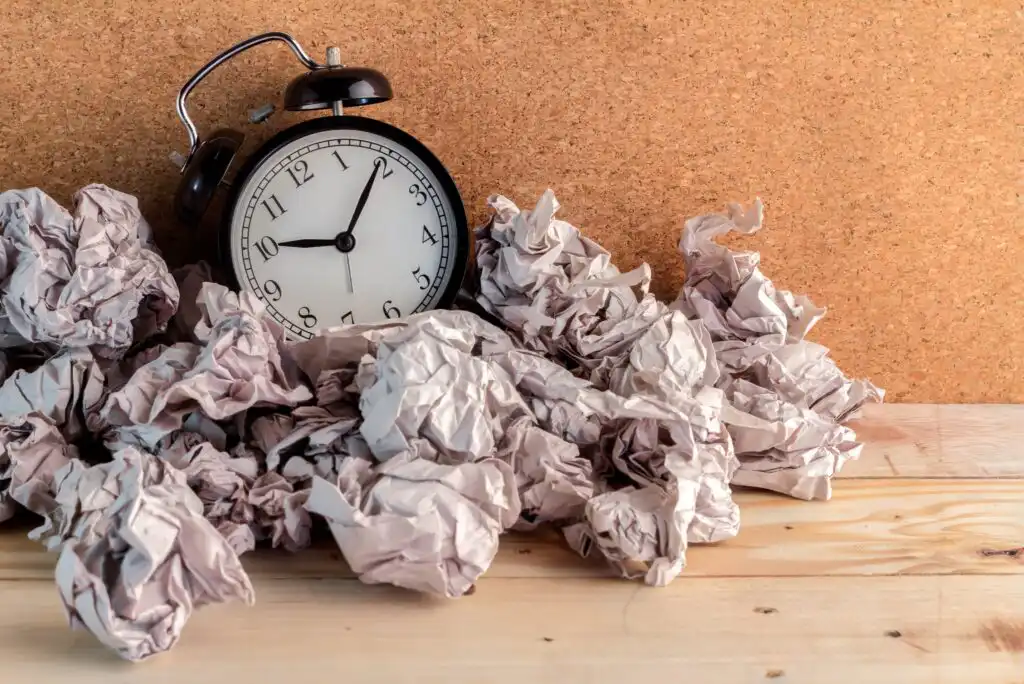 An analog alarm clock sits on a wooden surface surrounded by several crumpled sheets of paper, with a corkboard background.