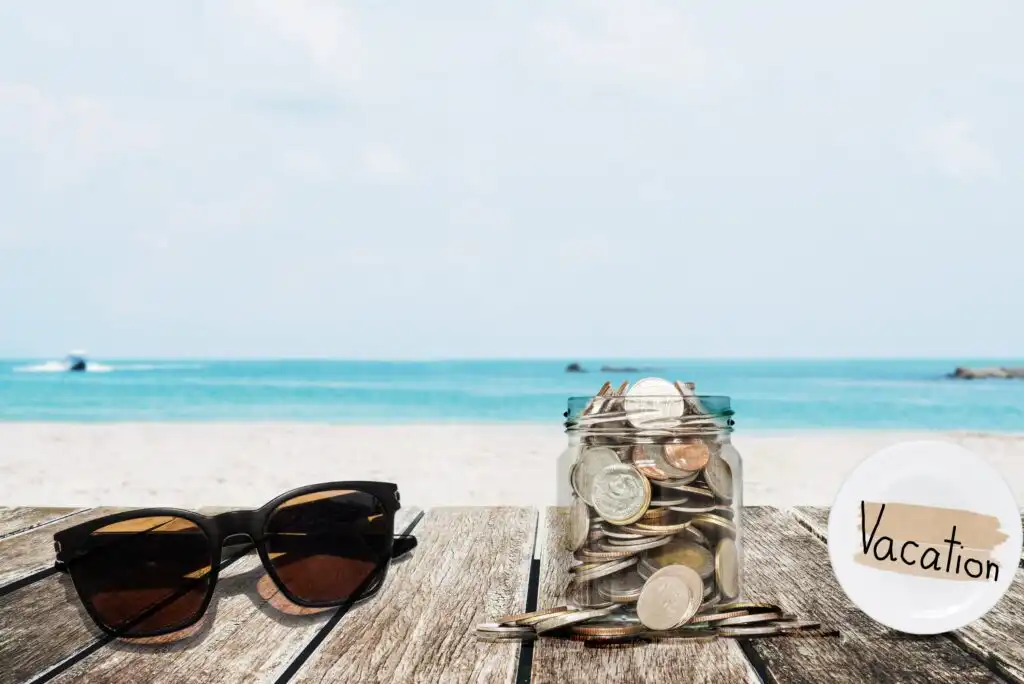 A pair of sunglasses and a jar filled with coins sit on a wooden table by the beach. The jar has a label that says “Vacation,” and the ocean and sky are visible in the background.
