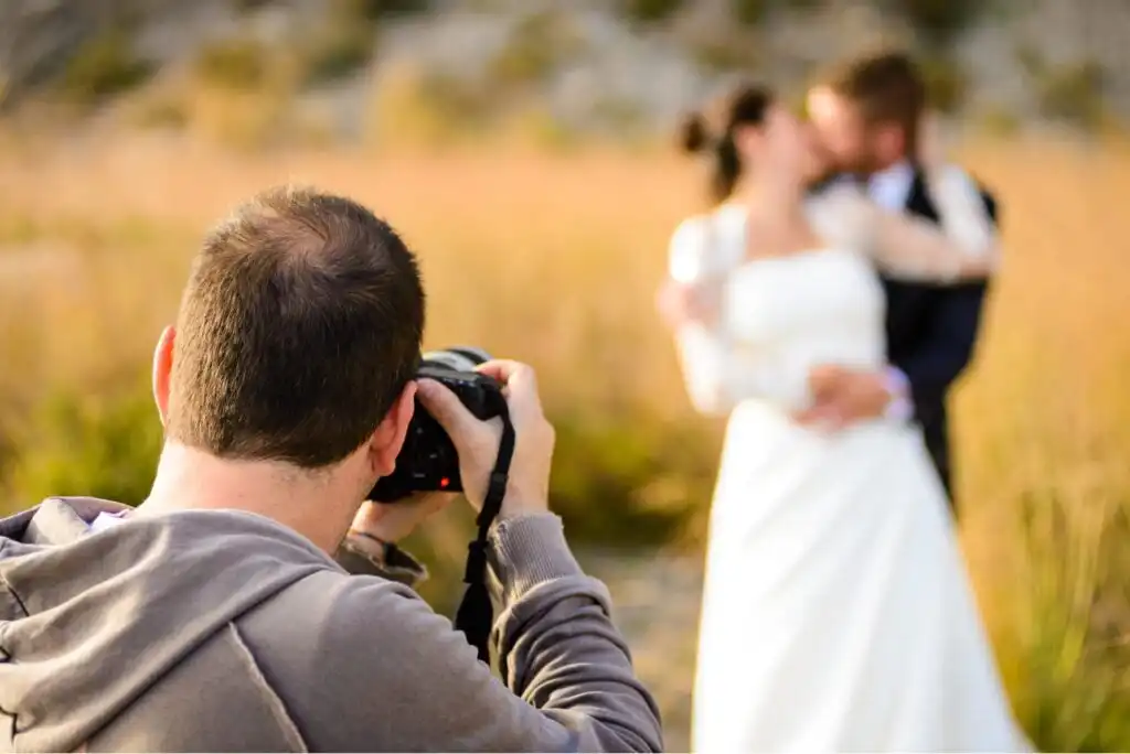 A photographer takes a picture of a bride and groom embracing and kissing outdoors, with blurred grass and trees in the background.