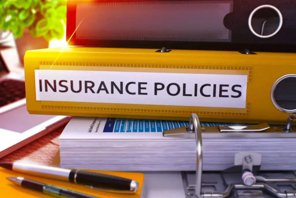 A yellow binder labeled Insurance Policies sits on a desk with paperwork and other binders, suggesting organization and management of insurance documents.