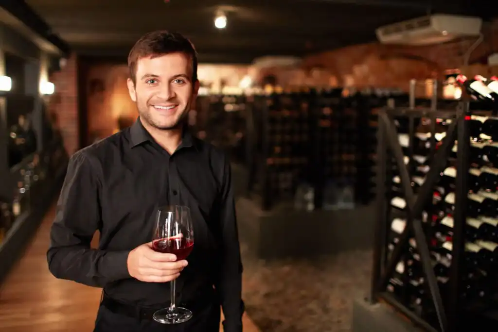 A smiling man in a black shirt holds a glass of red wine in a dimly lit wine cellar, with shelves of wine bottles visible in the background.