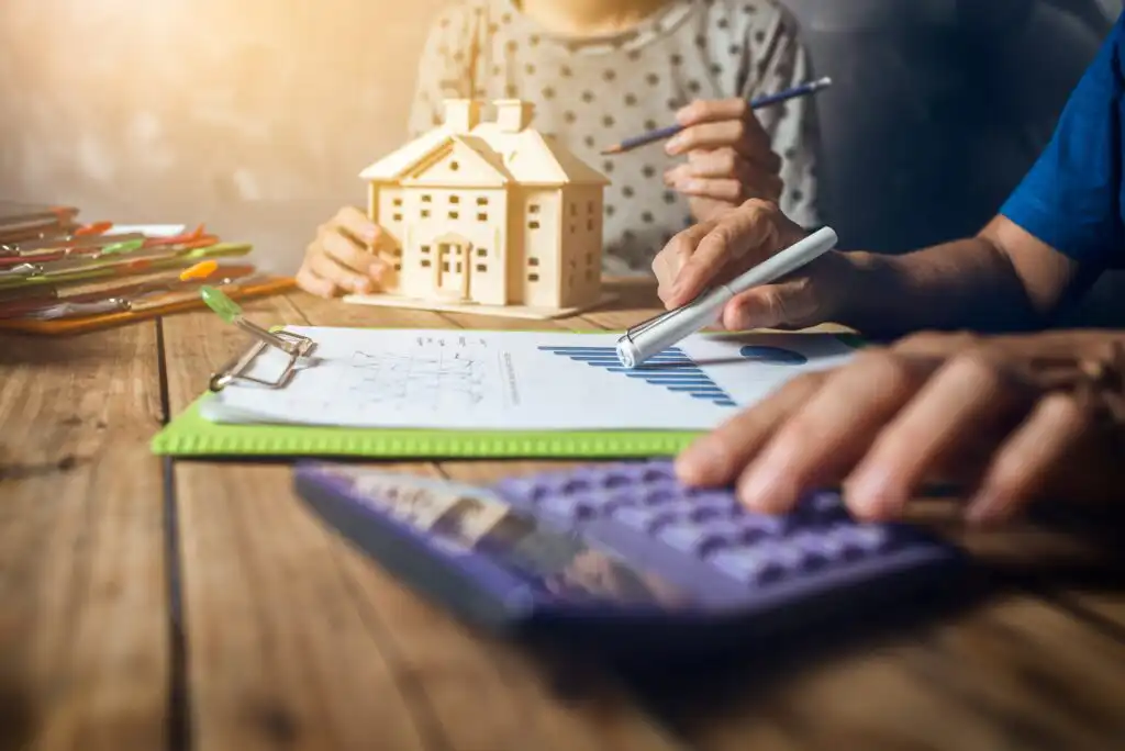 Two people sit at a wooden table with a small house model, charts on a clipboard, a calculator, and pens, discussing finances or planning a home purchase.