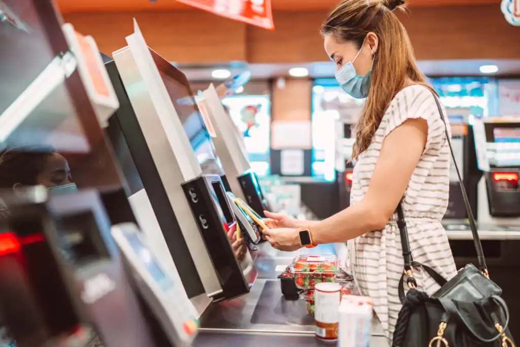 A woman wearing a face mask scans groceries at a self-checkout station in a store. She is holding a product and standing next to other items, including strawberries and bottled drinks.
