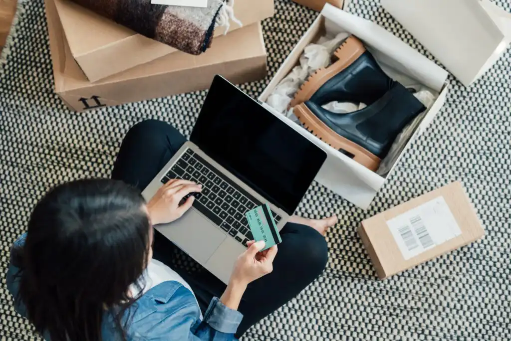 A person sits on the floor with a laptop, holding a credit card, surrounded by opened packages including a shoebox with black boots, shopping online.