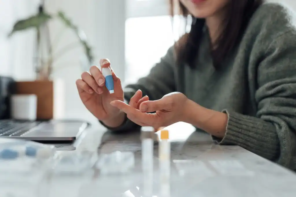 A woman sits at a table using a small device to prick her finger, likely for a blood test, with a laptop and test supplies nearby. The background is softly blurred.