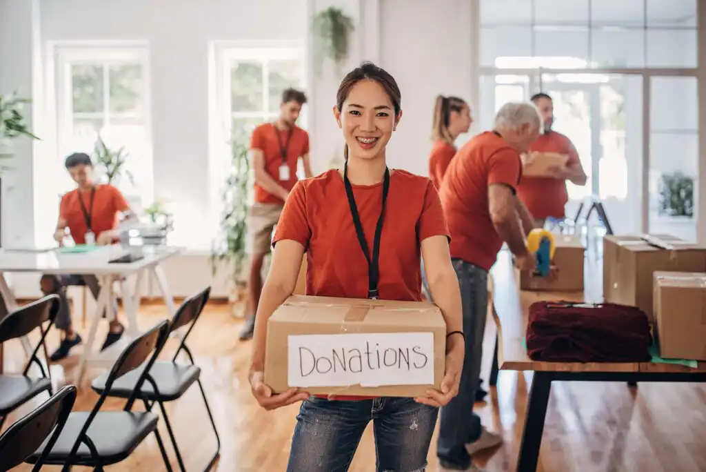 A smiling woman in a red shirt holds a box labeled Donations in a bright room, with other volunteers packing boxes and working in the background.