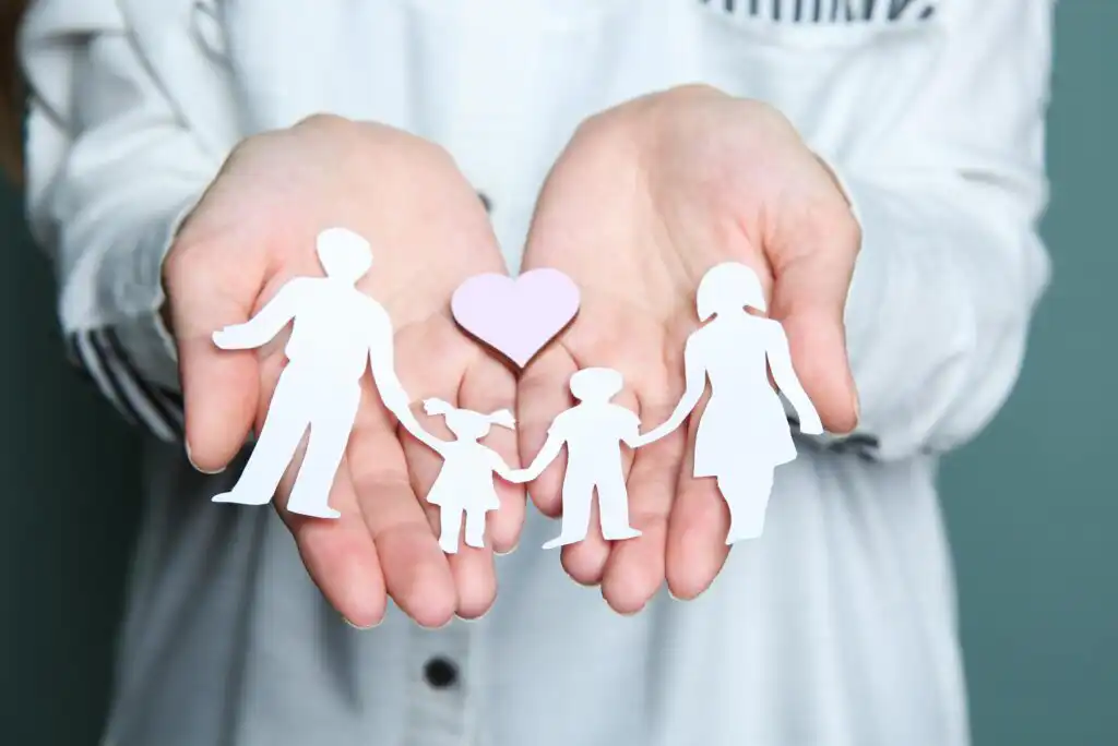 A person holds paper cutouts of a family—two adults and two children—along with a pink heart in their cupped hands, symbolizing care, love, and family support.